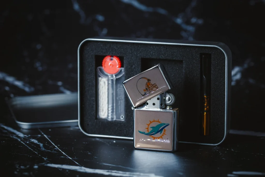 an image of a silver lighter on it with the Miami Dolphins mascot, with the optional gift box and accessories in the midground, on a black  and white background