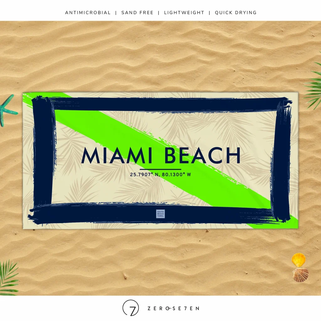 An image of a blue, tan, and green beach towel that says "miami Beach" with the GPS coordinates of the location on an artificial sand background. One of the best Miami gifts!