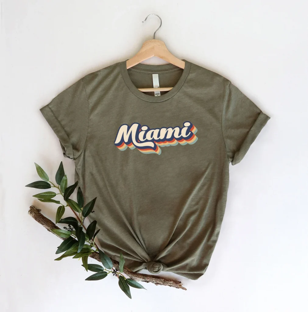 A picture of an olive green tshirt that reads "Miami" in a vintage rainbow font on a white background