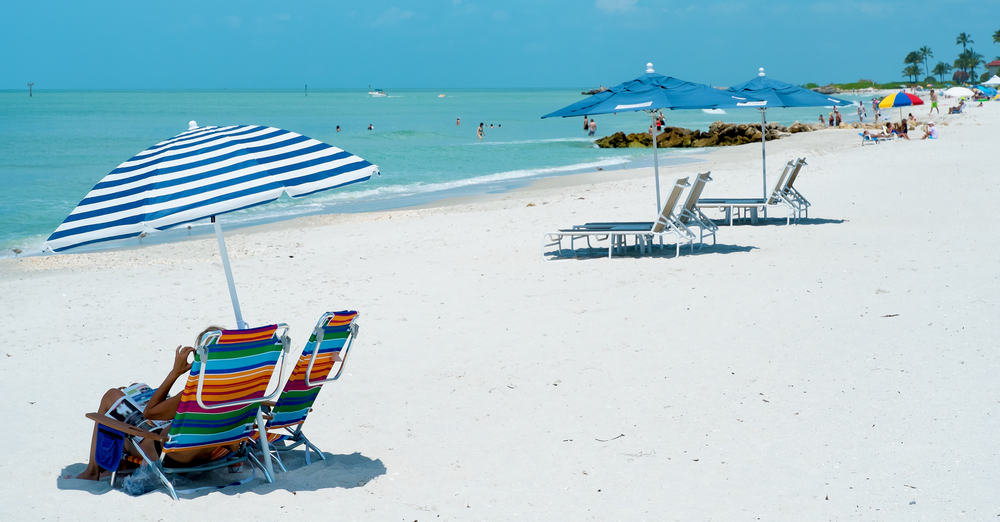 Beach chairs and umbrellas on a white sand beach in Naples Florida. You can see people swimming in the ocean and walking on the beach. 