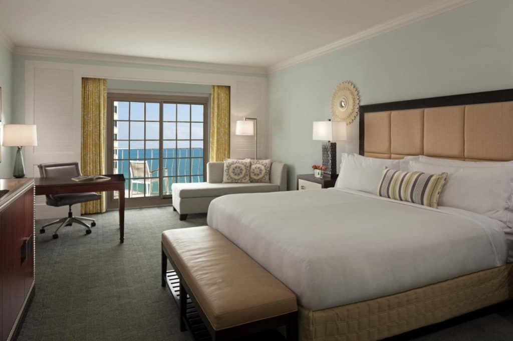 A hotel room with a king-sized bed, a small desk, a chaise lounge, and a sliding glass door that leads to a balcony with a view of the ocean. One of the best hotels on the beach in Naples Florida. 