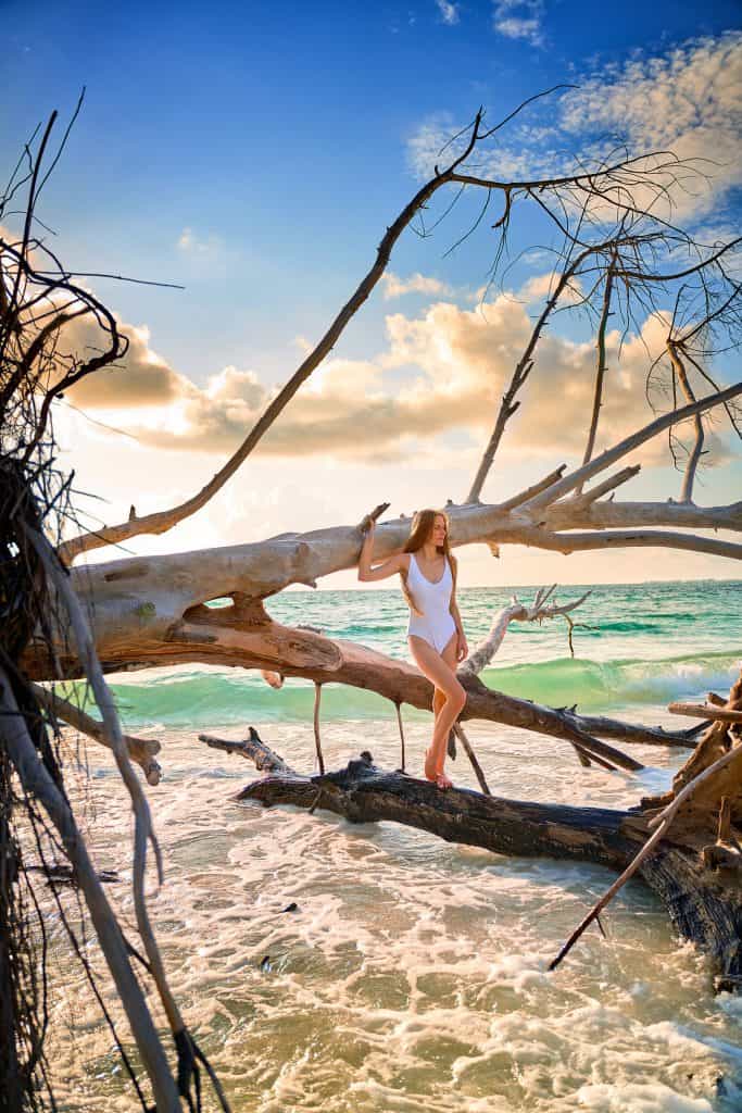 Girl in a white swimsuit standing against large pieces of driftwood in the water of Beer Can Island, one of the best beaches in Florida.