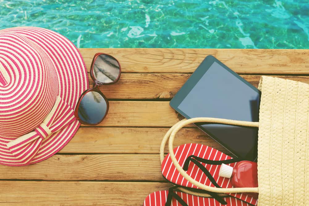 A bag spilling a hat, sunglasses, an e-reader, and other poolside essentials on a deck next to water.