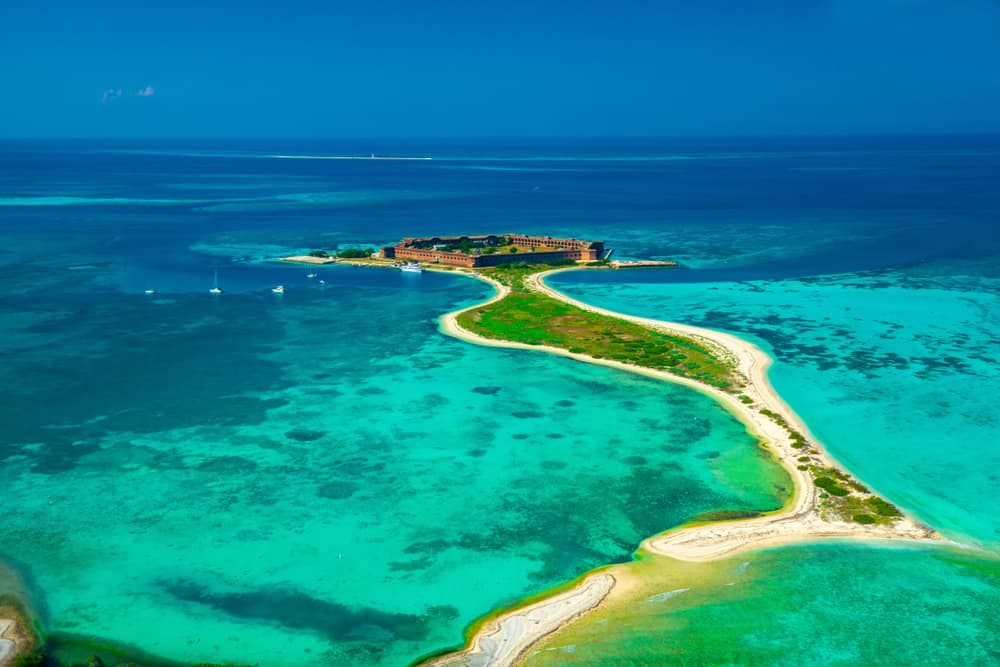 Aerial view of the Dry Tortugas National Park, in which a fort sits on an island surrounded by bright blue ocean water, with several sailboats nearby.
