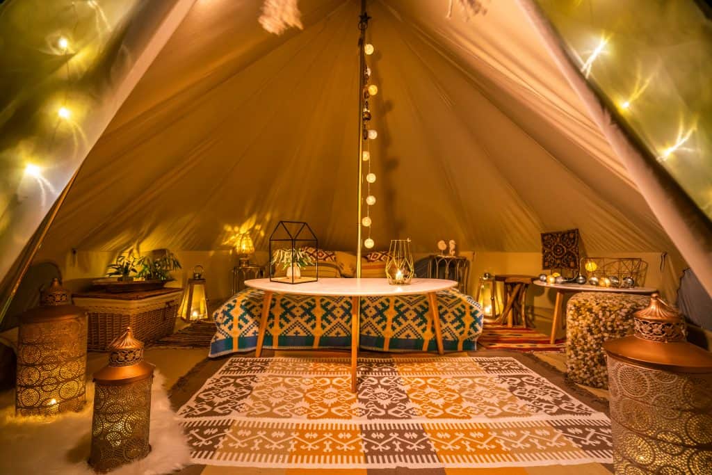 The interior of a glamping tent, including a bed, tables, and lanterns, and a decorative rug o the platform floor.