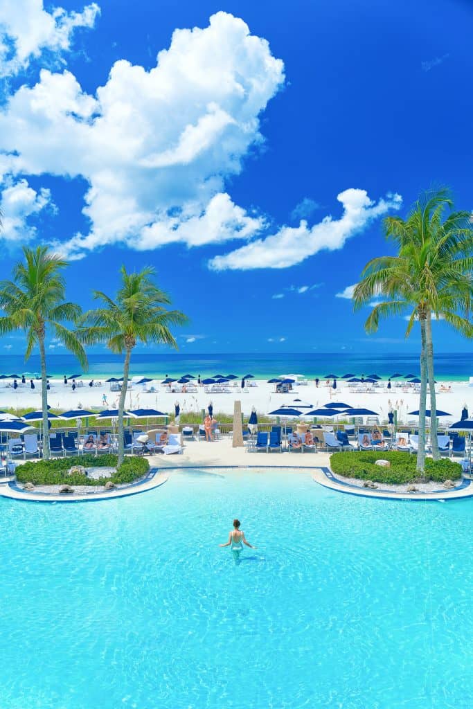 A woman with her back to the camera stands alone in a beachside pool, surrounded by blue umbrellas and lounge chairs at the Ritz-Carlton in Sarasota, FL, a great place for romantic getaways in Florida.