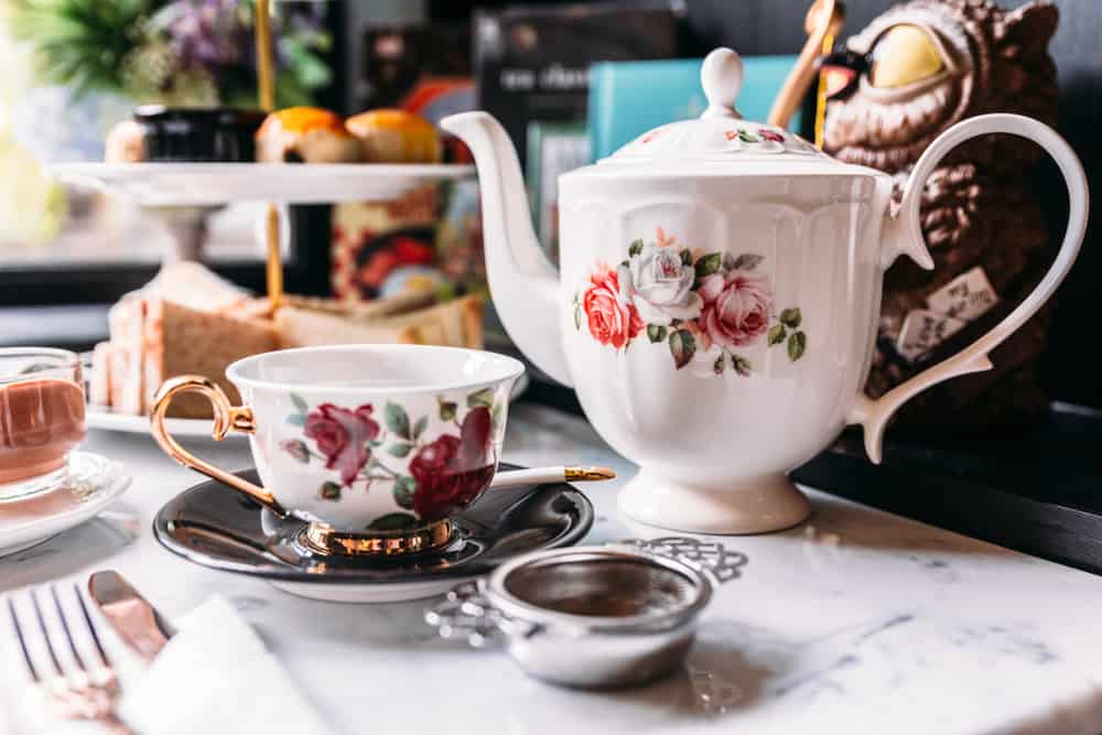 A pretty tea service with a rose painted teacup and tea pot and treats in the background.