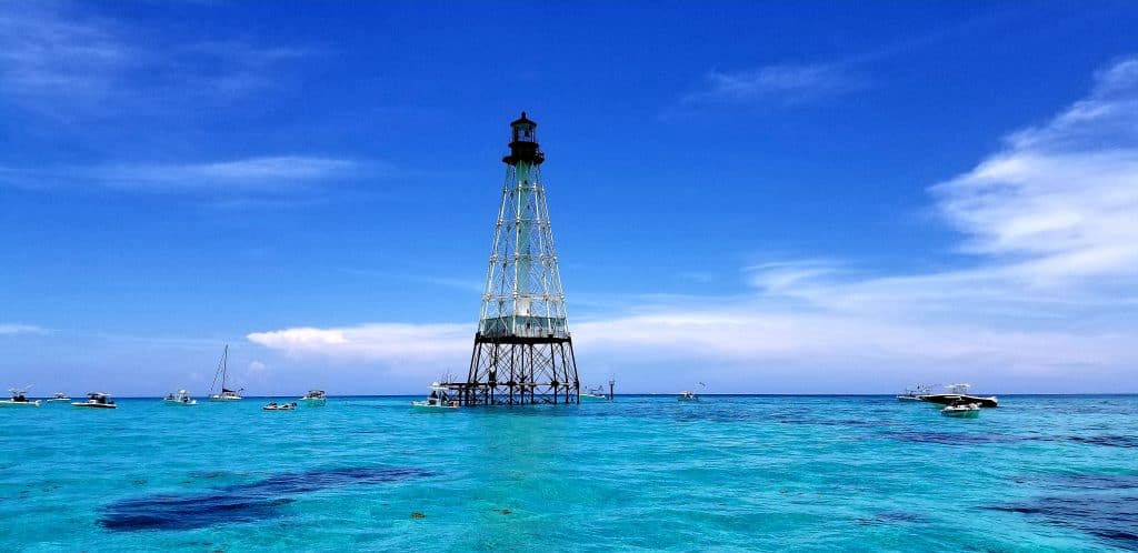 A distanced view of the Alligator Reef lighthouse, one of the best places to go in Islamorada, the foreground is the bright blue water, and the background is the lighthouse. There are boats in the distance and the sky is slightly cloudy