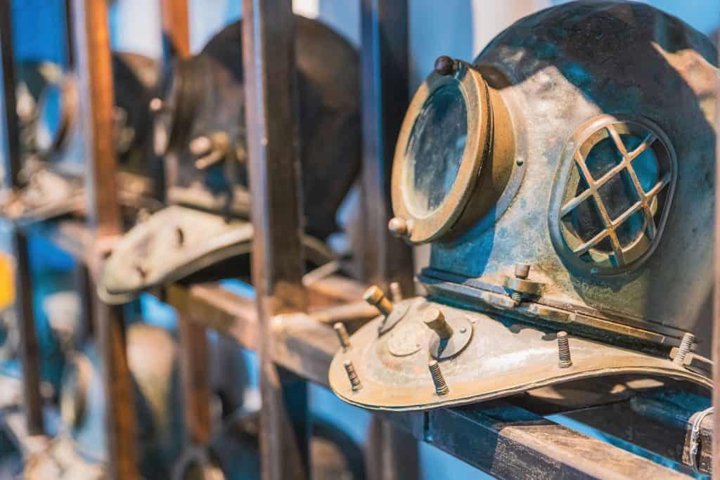 A picture of two antique diving helmets on a wooden shelf against a dark blue background. Just out of frame you can see there are more helmets on the shelves 