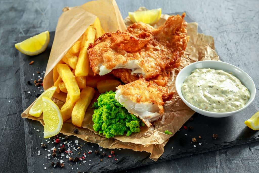 A picture of battered fish and fries with a side of tartar sauce and lemon wedges against a dark grey stone background. The food is appetizing and colorful
