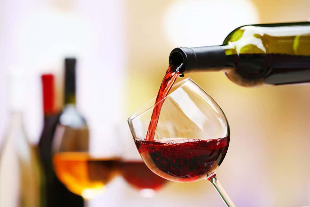 A picture of a glass of red wine being poured from a green bottle, there are other colorful bottles out of focus in the background 