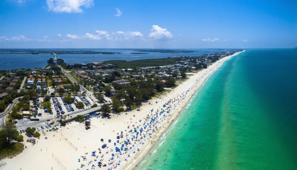 An aerial view of Bean Point Beach in Anna Maria Island, the color palette is dark green from the trees growing on the island, the white sand, and gorgeous green and blue waters. Colorful umbrellas dot the white sand creating a colorful scene
