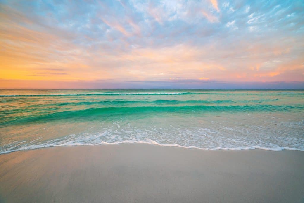 A straight on point of view photo of the ocean at Destin Beach, right as the sun is rising, creating a color palette of white sand, emerald water, and pastels from the sunrise