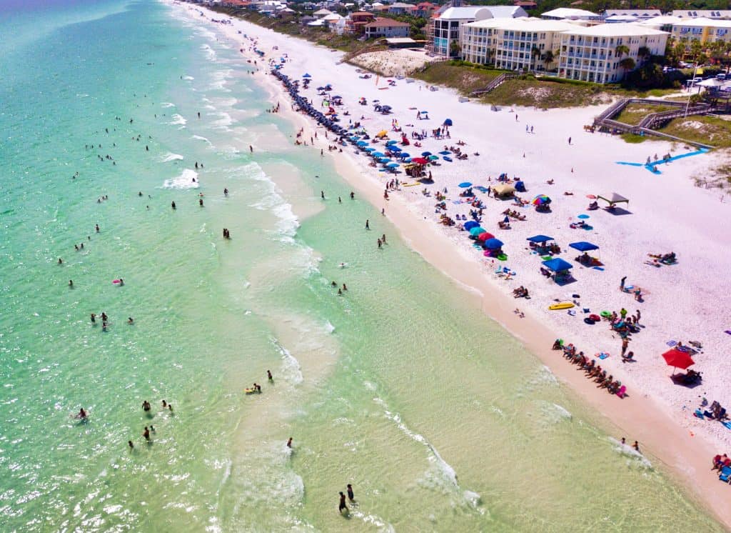 An aerial view of Santa Rosa, beach there is a long line of dark blue umbrellas hugging the shoreline with a few odd red and green ones too. There are lots of people in the water but it is not crowded, and the waves make the ocean water seem extra textured