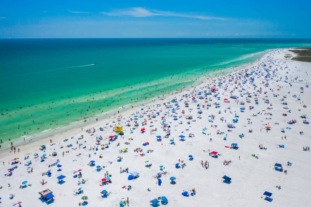 An aerieal view of Siesta key, one of the best white sand beaches in Florida, showings a rainbow of different colored umbrellas, a colorful yellow lifeguard house, and the emerald waters