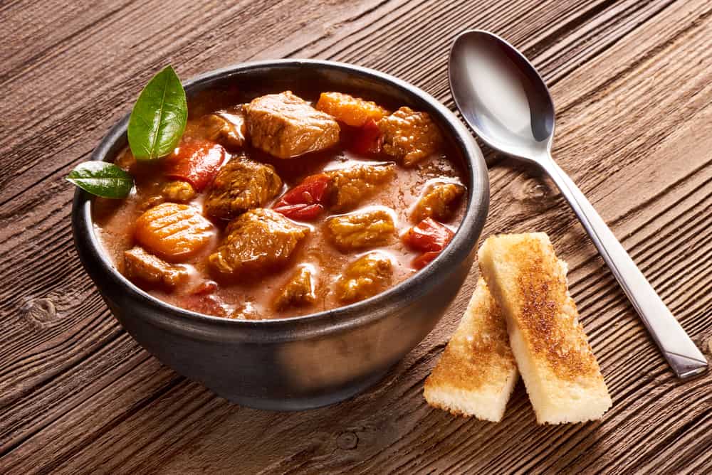 A heaping bowl of goulash with meat and veggies on a wooden background.