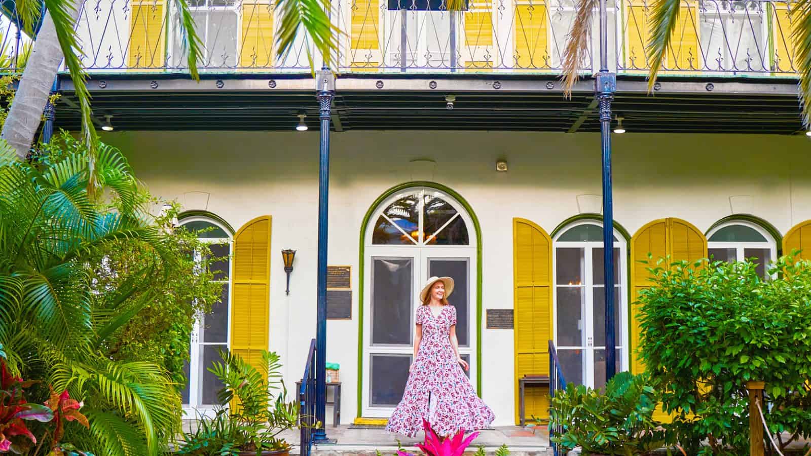 A woman in a dress and sun hat smiles on the front steps of the Ernest Hemingway Home and Museum.