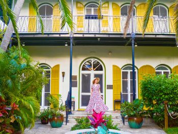 A woman in a dress and sun hat smiles on the front steps of the Ernest Hemingway Home and Museum.
