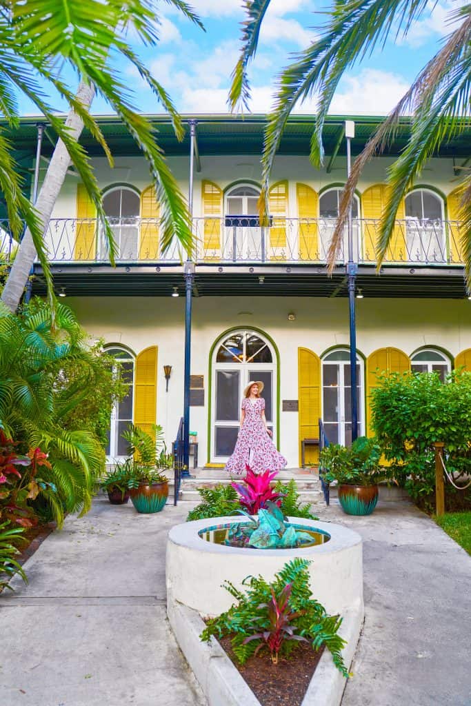 A woman in a dress and sun hat smiles in front of yellow shuttered windows on the front steps of the Ernest Hemingway Home and Museum.