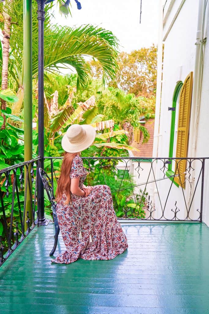A woman in a long floral dress and sun hat sits on a metal chair, looking away, on the porch of the Ernest Hemingway Home.