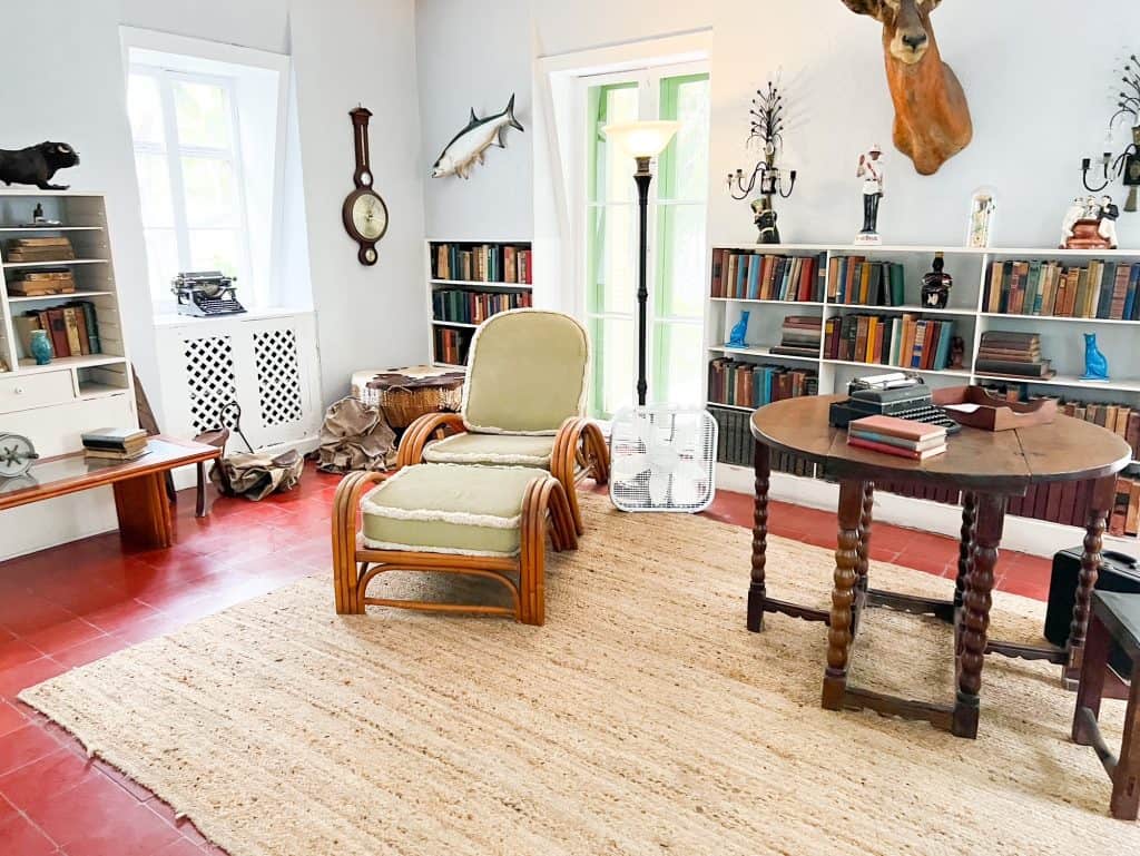 A two-piece chair, and a wooden table with a typewriter sit surrounded by books in the author's writing studio at the Ernest Hemingway Home and Museum.