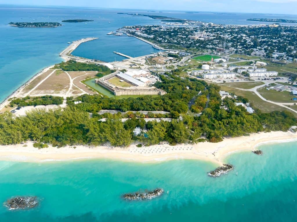 Aerial view of Fort Zachary Taylor State Park with the beach in the foreground and the four stone walls of the fort in the background, surrounded by foliage.