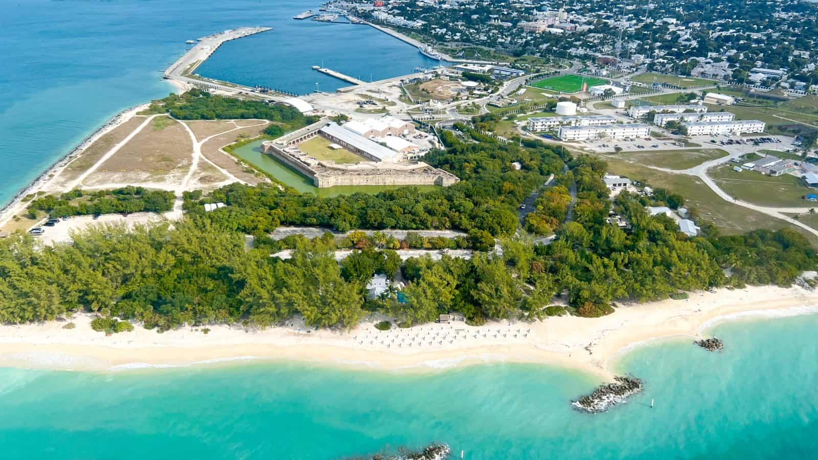 Aerial view of Fort Zachary Taylor State Park with the beach in the foreground and the four stone walls of the fort in the background, surrounded by foliage.