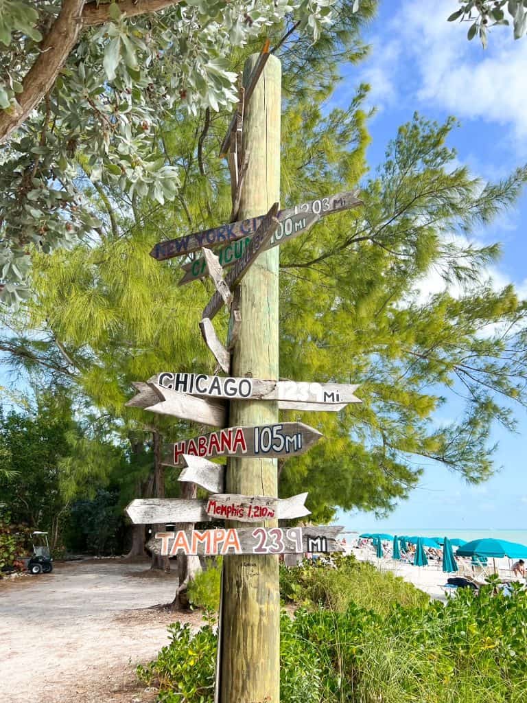 A collection of arrow-shaped wooden signs on a telephone pole near the beach, with different city names painted on them and how many miles they are from Fort Zachary Taylor State Park.