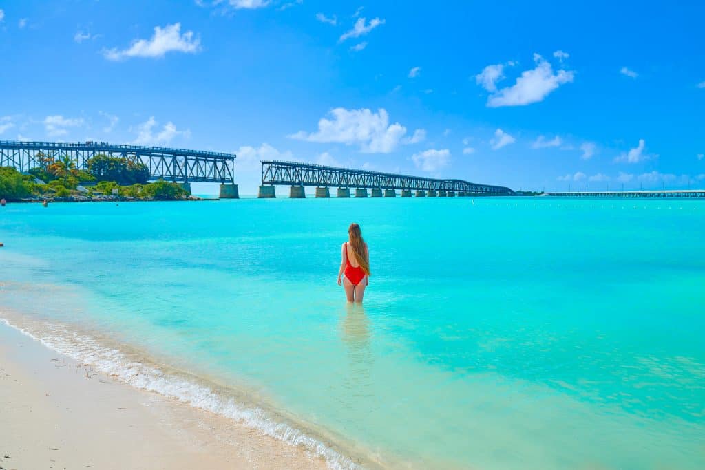 Victoria faces and walks toward Old Bahia Bridge in a red bathing suit, wading in the clear blue water at Bahia Honda State Park.