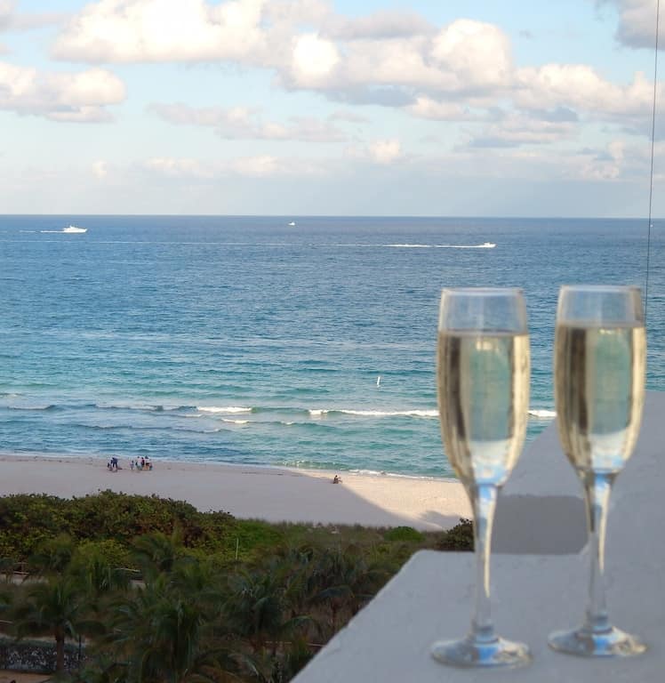 closeup of two champagne flutes on the balcony overlooking a white sandy beach and strikingly blue water. 