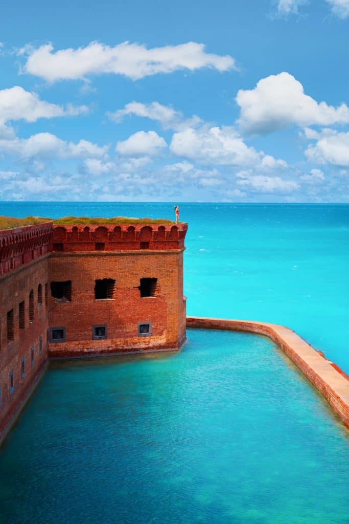 a girl in a red swimsuit standing on top of the fort overlooking the crystal clear turquoise water