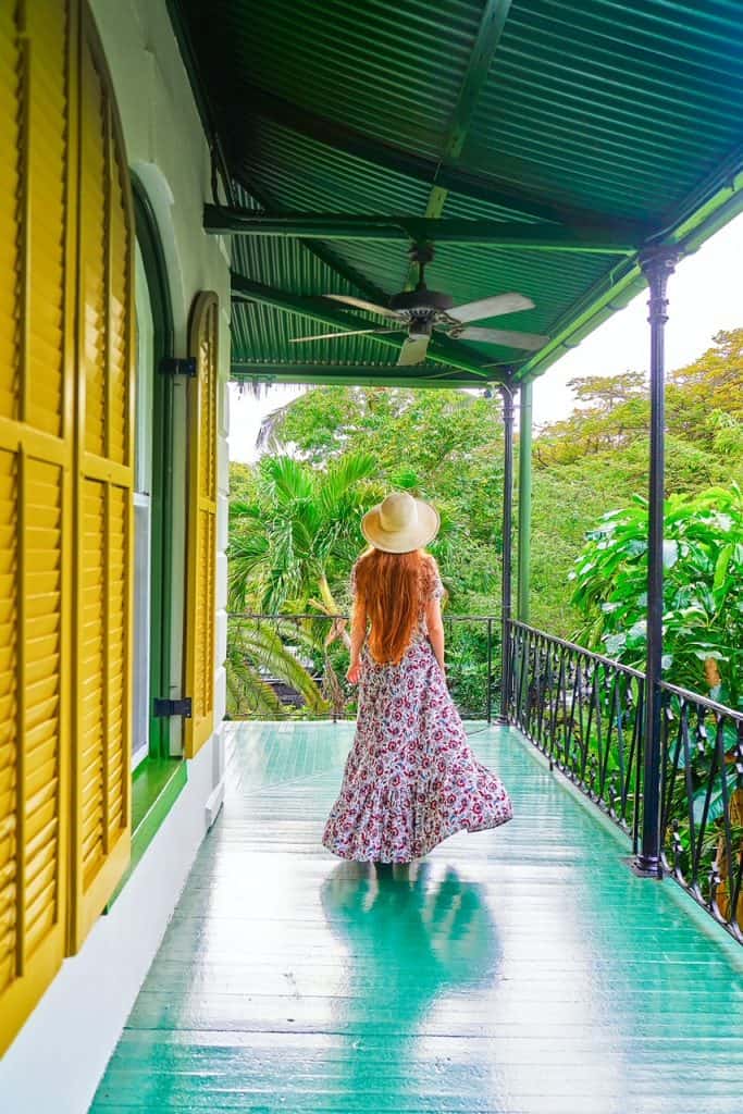 A girl in a flower flower dress with a. hat on a green balcony overlooking lush foliage with yellow shutters