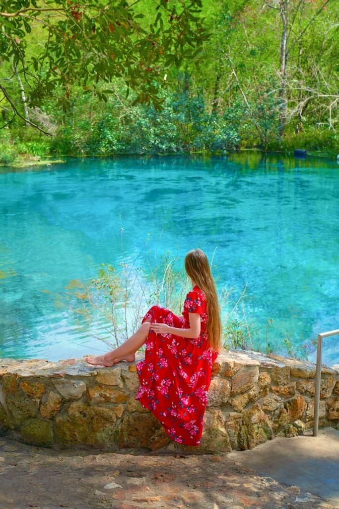 A woman in a red floral dress perches herself on the edge of a stone wall and looks over the blue water at Ichetucknee Springs.