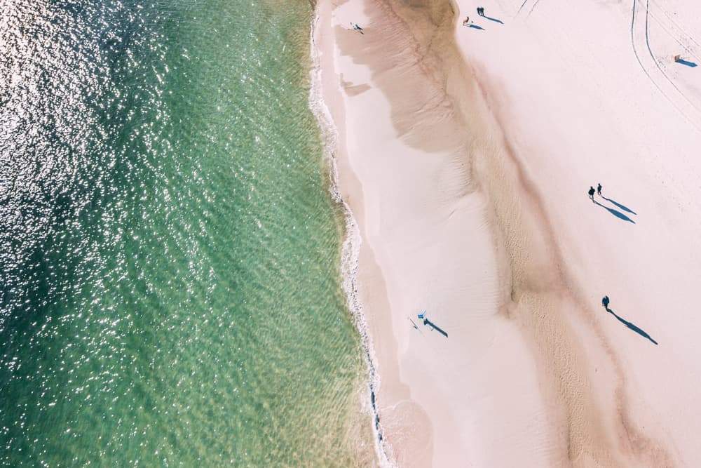 An aerial shot of one of the best Panhandle beaches in FL, with emerald-colored ocean on the left, and several people walking along clean sand on the right.