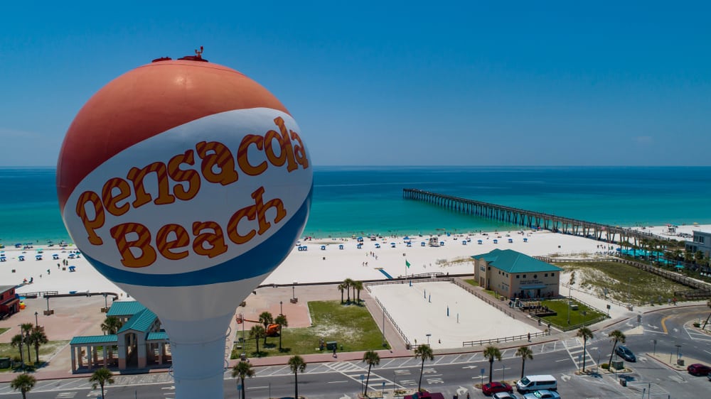 The Pensacola Beach water tower, painted like a beach ball, stands in the foreground with one of the best Panhandle beaches in the background, with a fishing pier stretching out into blue water on a sunny day in FL.