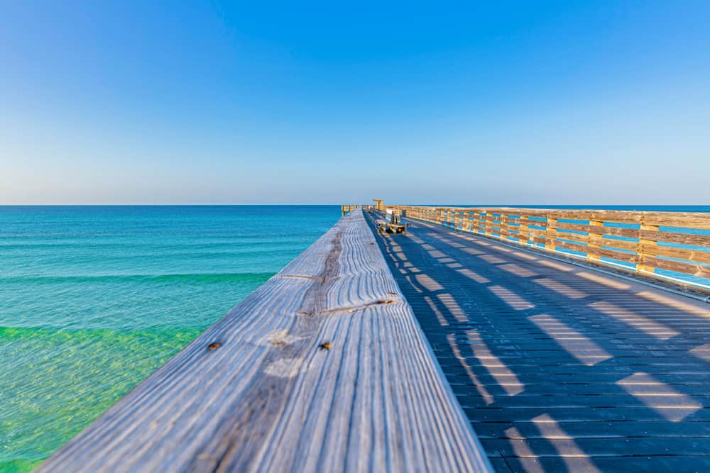 Shadows fall on the wooden railing of the pier at St. Andrews State Park during sunset over blue water.