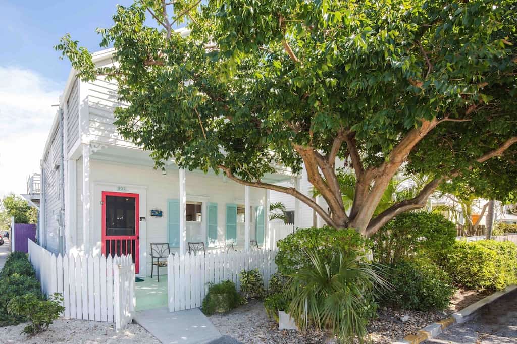 The whitewashed exterior of the Old Town Cottage. The cottage has a white picket fence, teal shutters, a coral door, and lush landscaping. 