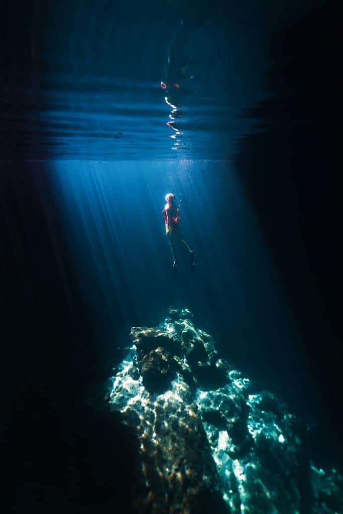 A swimmer approaches the surface of the water at Devin's Den: her flippers propel her forward and the rays of light catch her red bathing suit in the water.