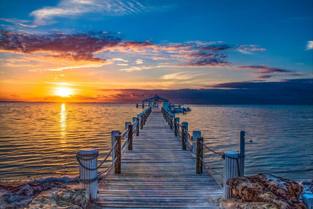 A stunning shot looking out at a dock during sunrise in islamorada, walking out to the edge will have you feeling like you're out in the middle of the ocean. The dock has rope and pillar railing on either side, and at the far end of the dock there is a small gazebo