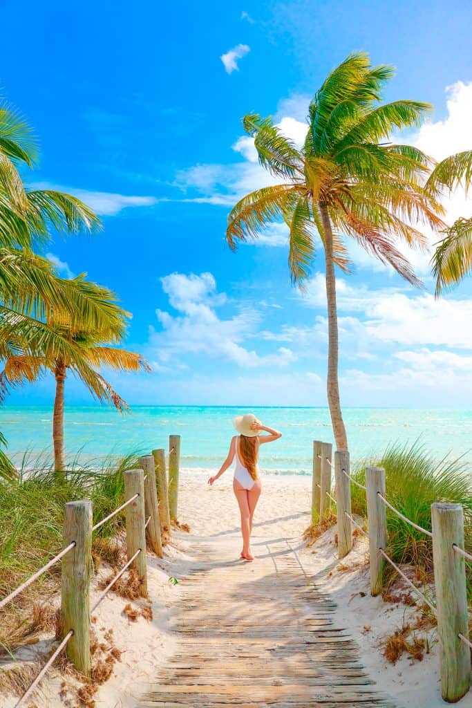 A shot of a beautiful woman in a white bathing suit wearing a large hat, she is facing the ocean's bright blue and green waters, framed by palm trees on either side, standing on a path that has lead her there