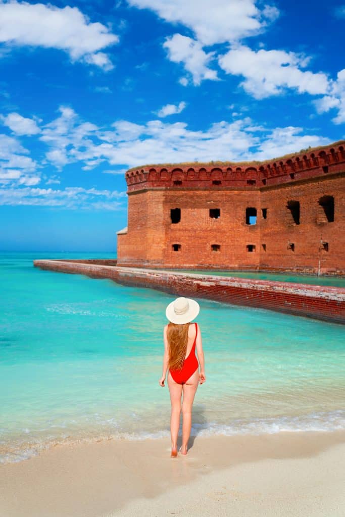 A photo of a beautiful woman wearing a red bathing suit and a big white hat, standing on white sand. In the background, the red and orange brick of the fort at Dry Tortugas provides a beautiful contrast in color