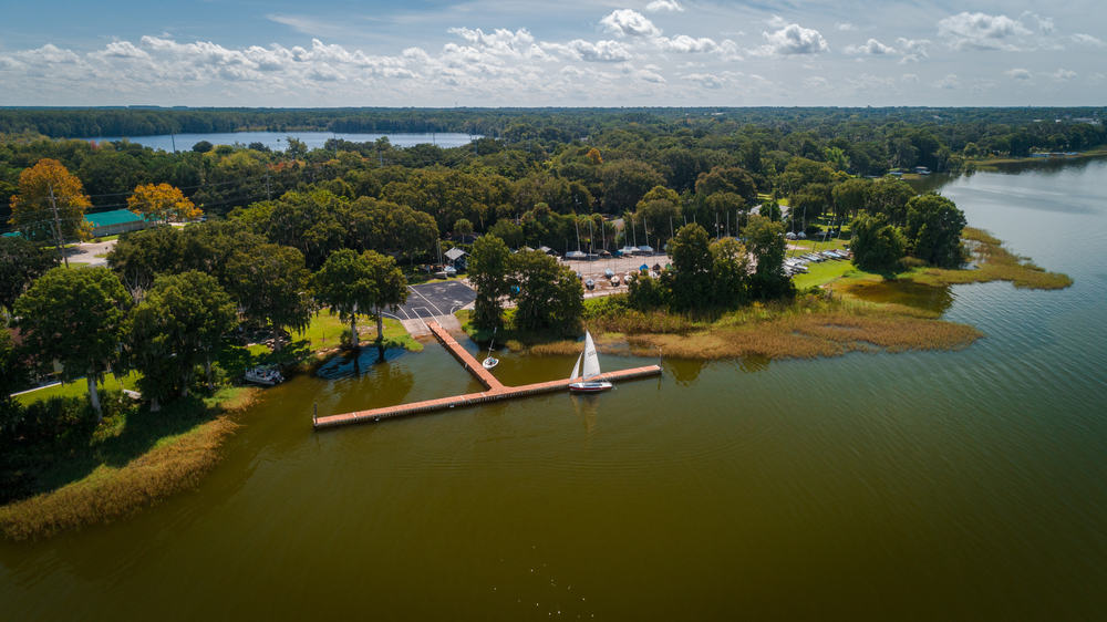 Aerial view over Lake Eustis Sailing Club. A sailing boat in my the dock and there are trees and a car park in the background. 