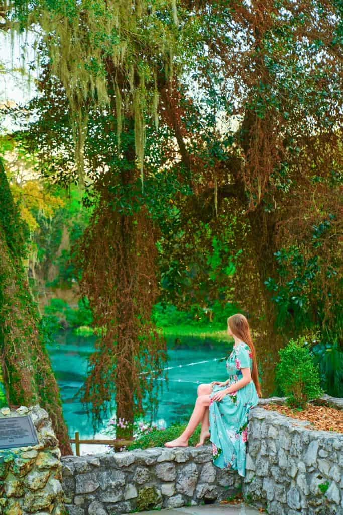 A woman finds a look out and gazes out over the river at Rainbow Springs. This lookout is a niche area that has stone walls, and is full of moss, but still offers views of the blue water.