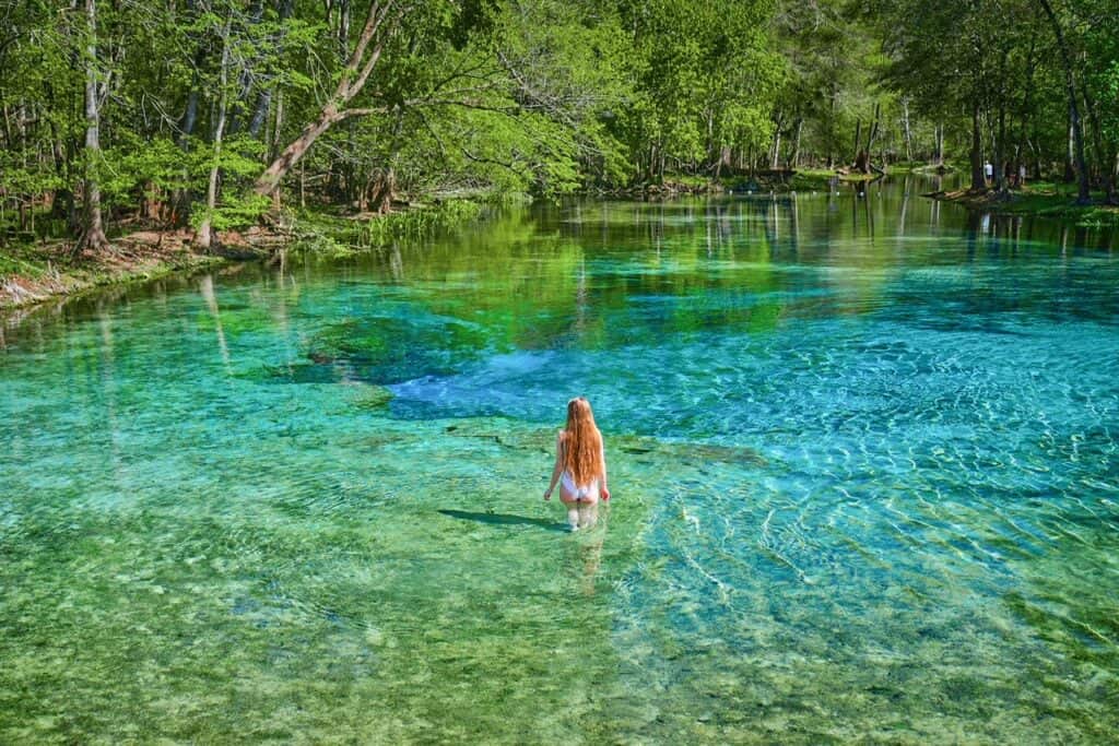 A woman with long hair walks out into the bright blue waters of a spring.