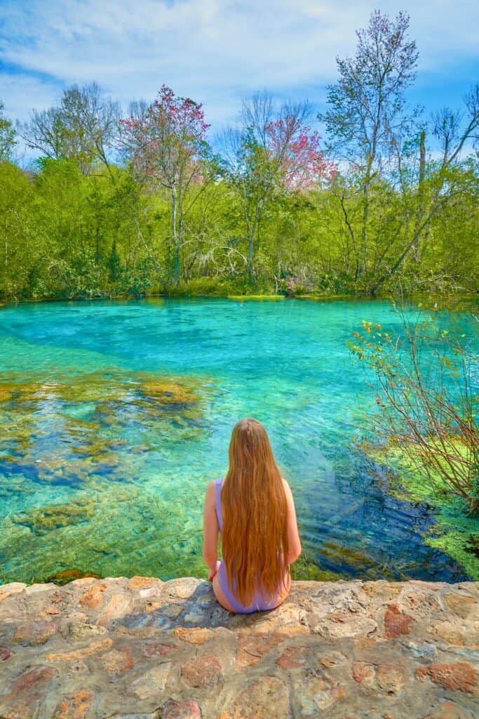 A woman with long loose hair in a lavender swim suit sits on the rocky edge of colorful Ichetucknee Springs.