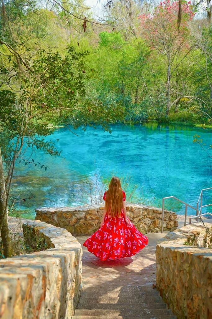 A woman in a red, floral dress stands on the stone pathway near the bright blue Ichetucknee Springs.