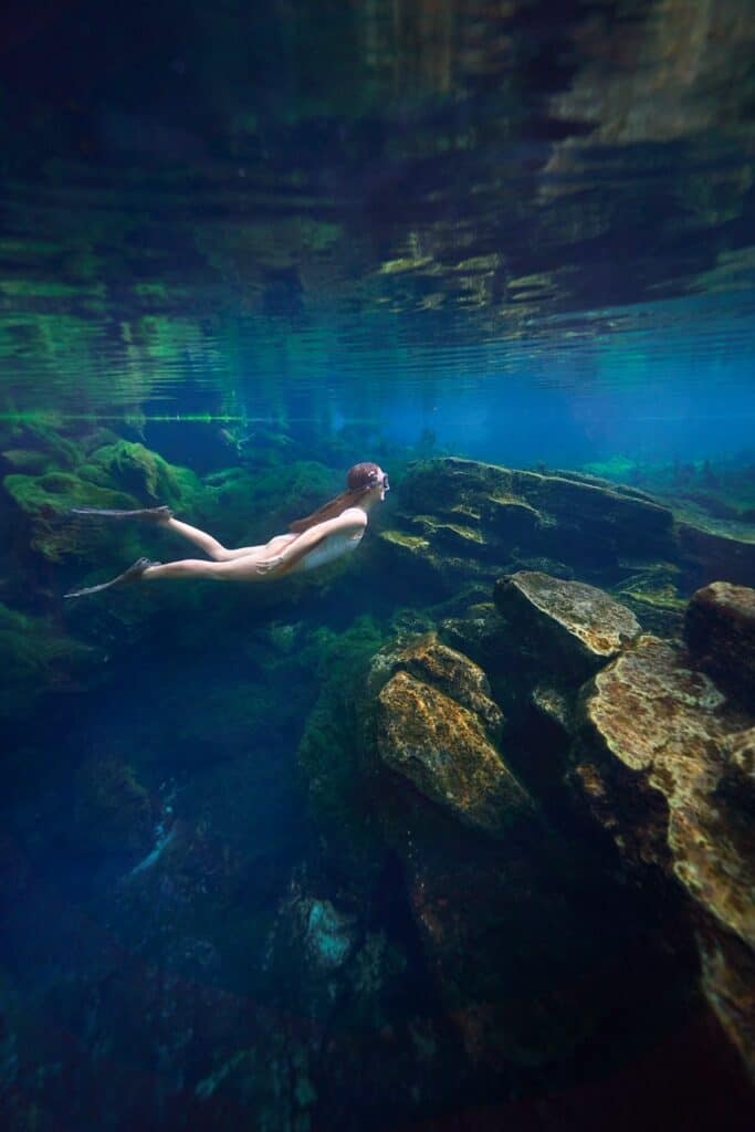 Woman in white swimsuit and snorkel gear swims underwater over rocks in Peacock Springs.