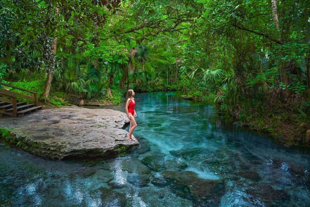 A woman in a red swim suit standing on a rock on the edge of the beautiful blue water of Rock Springs under a canopy of green.