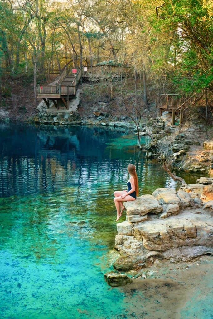 A woman in a black swim suit sits on a rocky ledge over the water of Royal Spring with the jumping platform in the background.