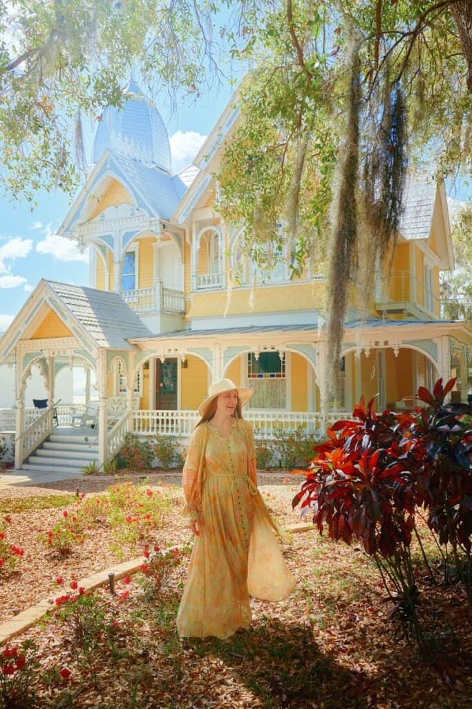 A woman in a yellow sundress and hat stands smiling in front of a yellow and white Victorian home in Mount Dora, Florida.